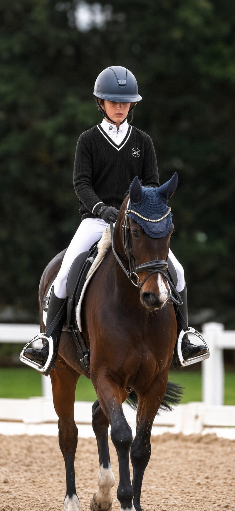A rider in equestrian competition attire sits on a dark brown horse in an outdoor arena. The rider wears a navy helmet, a black sweater with white trim, white breeches, and black boots.