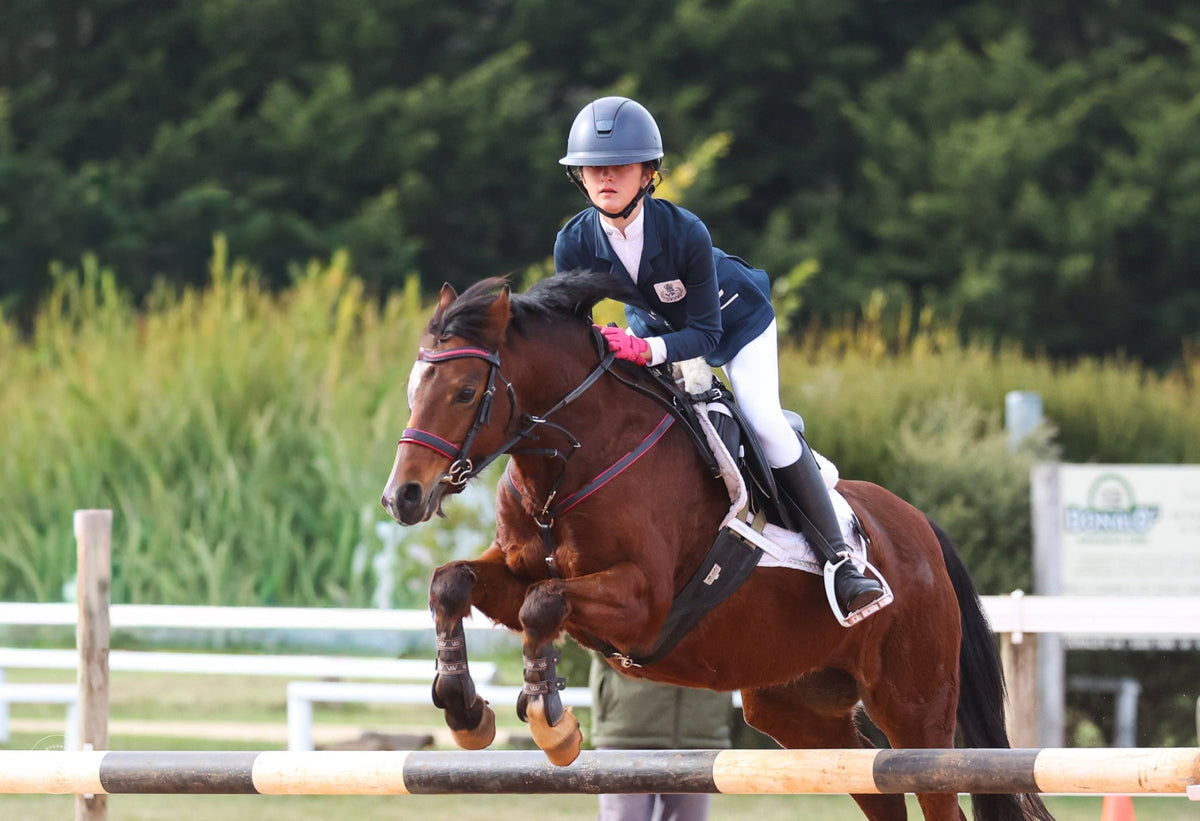 Young rider in a navy jacket, white breeches and black riding boots jumping a bay horse over an arena rail