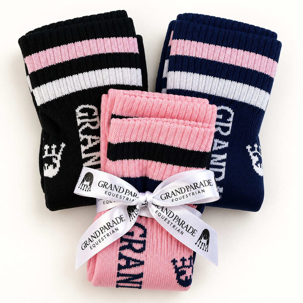 Three pairs of folded crew socks arranged together on a white background: one black pair with pink and white stripes, one navy pair with pink and white stripes, and one pink pair with dark navy stripes. A white ribbon bow tied around the center pair reads GRAND PARADE EQUESTRIAN.