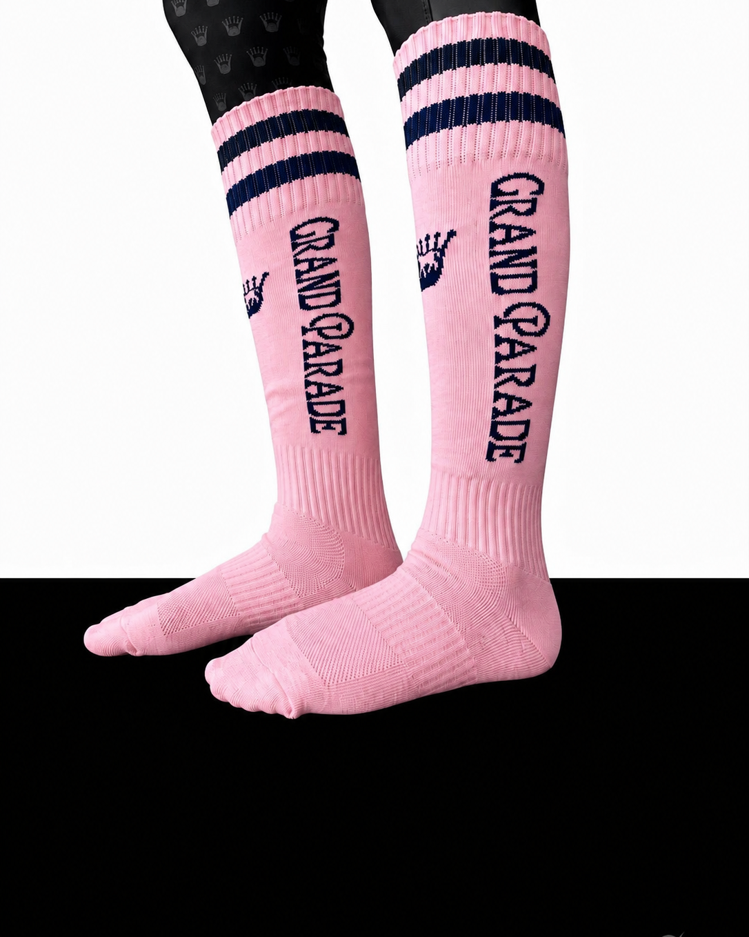 Pink crew socks with navy blue stripes and 'GRAND PARADE' text, shown on a model's legs against a split white and black background.