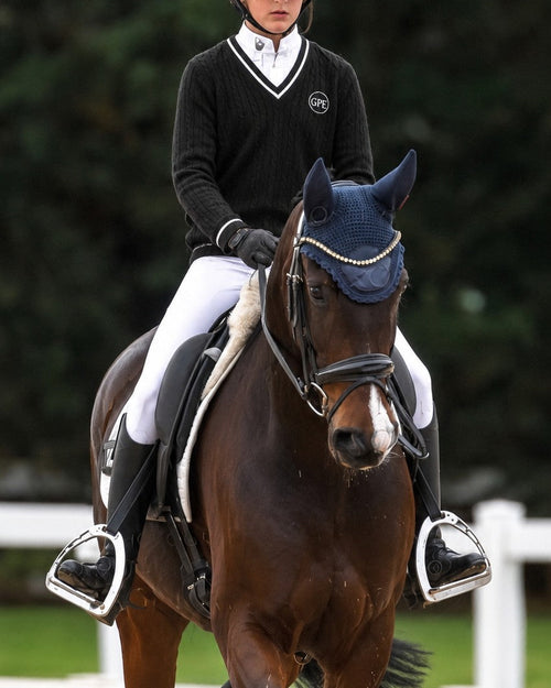 A rider in equestrian competition attire sits on a dark brown horse in an outdoor arena. The rider wears a navy helmet, a black sweater with white trim, white breeches, and black boots.