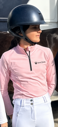 A person wearing a black riding helmet and a light pink long-sleeve equestrian quarter-zip shirt with a visible 'Grand Parade Equestrian' logo, paired with white Pro Breeches.
