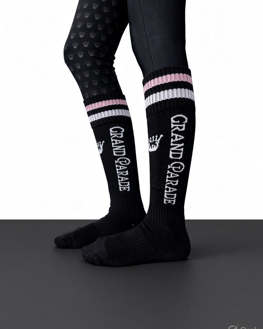 Black knee-high athletic socks with white and pink striped cuffs, featuring the words 'GRAND PARADE' in white, shown on a model's legs against a white and dark gray background.