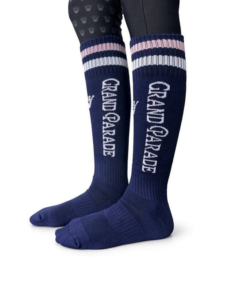 A pair of navy blue knee-high riding socks with white and light pink striped cuffs, featuring 'GRAND PARADE' printed vertically in white on the lower leg, shown on a model's legs against a white background.