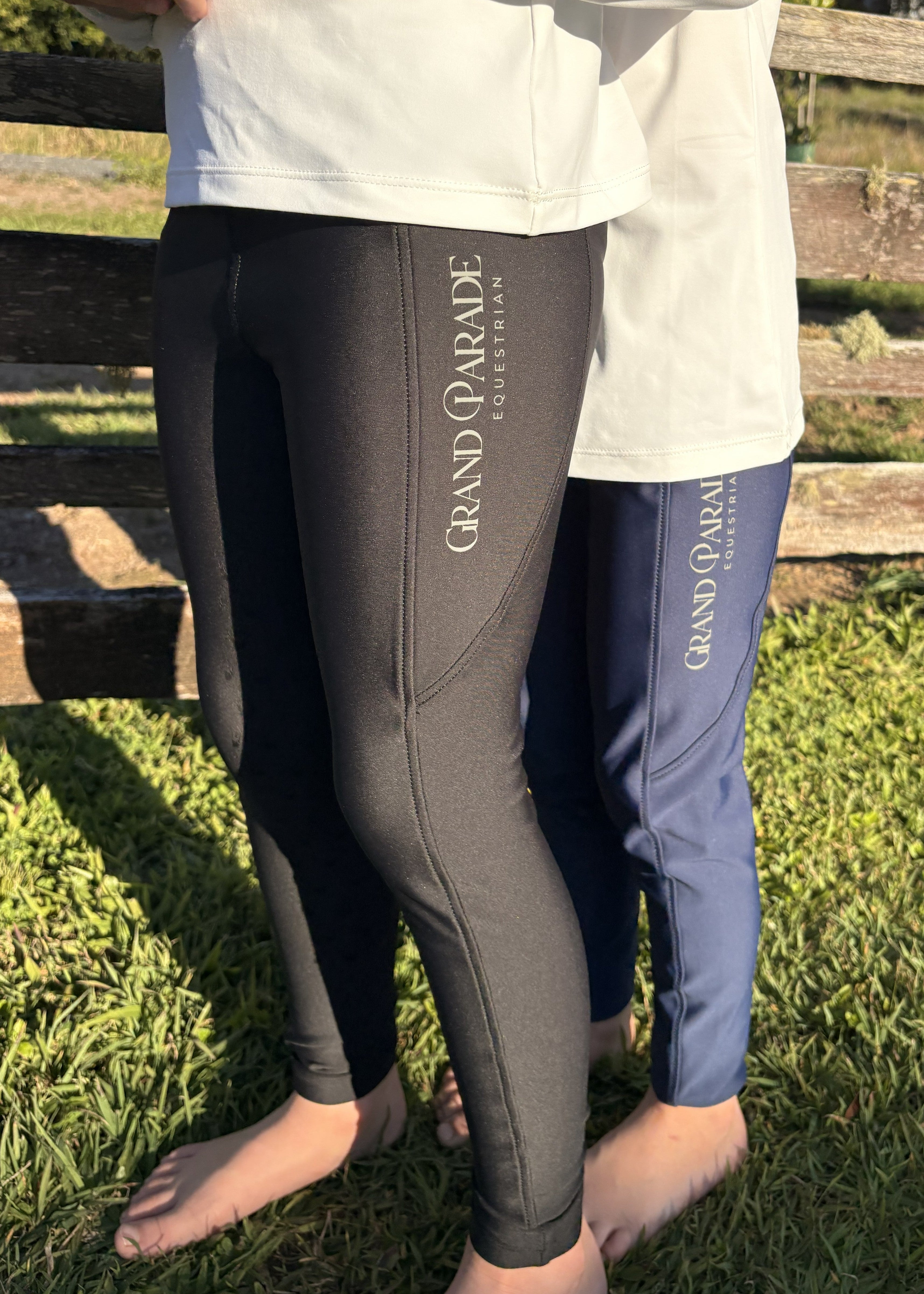Two people standing barefoot outdoors on grass in front of a wooden fence, wearing GRAND PARADE EQUEATRIAN brand leggings. One person wears black leggings and the other wears navy blue leggings, both with white long-sleeve tops partially visible.
