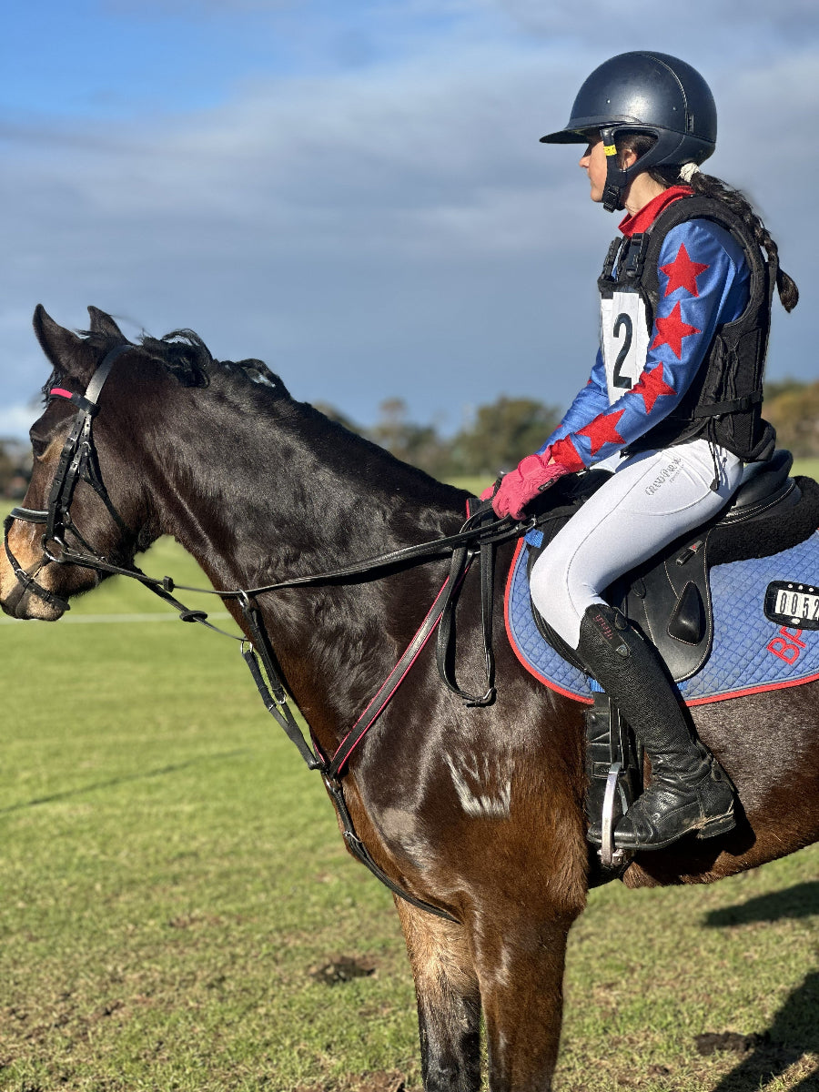 A person riding a dark brown horse outdoors on a grassy field under a blue sky. The rider wears a navy helmet, a blue and red patterned long-sleeve shirt on behalf of Bullengarook Pony Club, white breeches, and black riding boots.