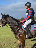A person riding a dark brown horse outdoors on a grassy field under a blue sky. The rider wears a navy helmet, a blue and red patterned long-sleeve shirt on behalf of Bullengarook Pony Club, white breeches, and black riding boots.