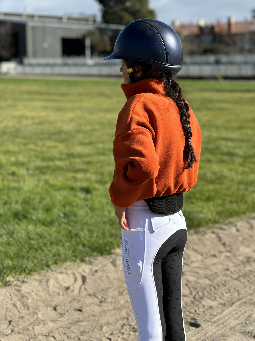 A child equestrian standing in profile on a sandy track wearing a navy riding helmet, an orange fleece jacket, white breeches with black knee panels, and a black belt, with a green grass field in the background.