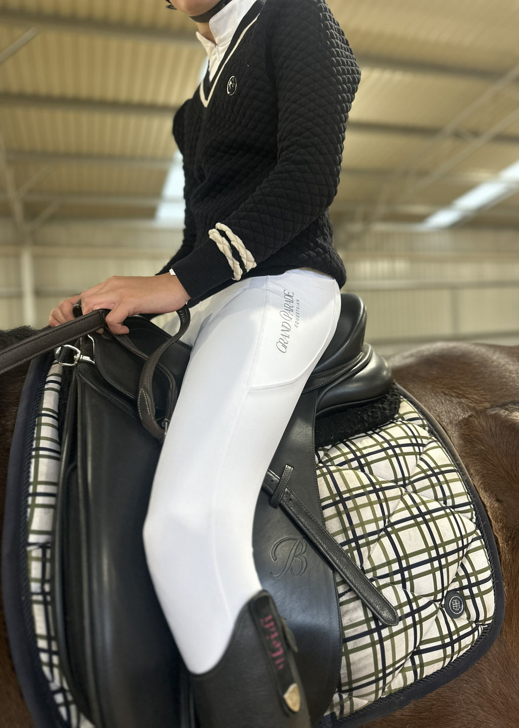 A person in equestrian attire, wearing a black knit sweater and white riding breeches with 'Grand Parade Equestrian' printed on the thigh, seated on a horse with a black leather saddle and a cream-and-black plaid saddle pad inside an indoor riding arena.