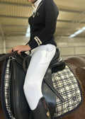A person in equestrian attire, wearing a black knit sweater and white riding breeches with 'Grand Parade Equestrian' printed on the thigh, seated on a horse with a black leather saddle and a cream-and-black plaid saddle pad inside an indoor riding arena.