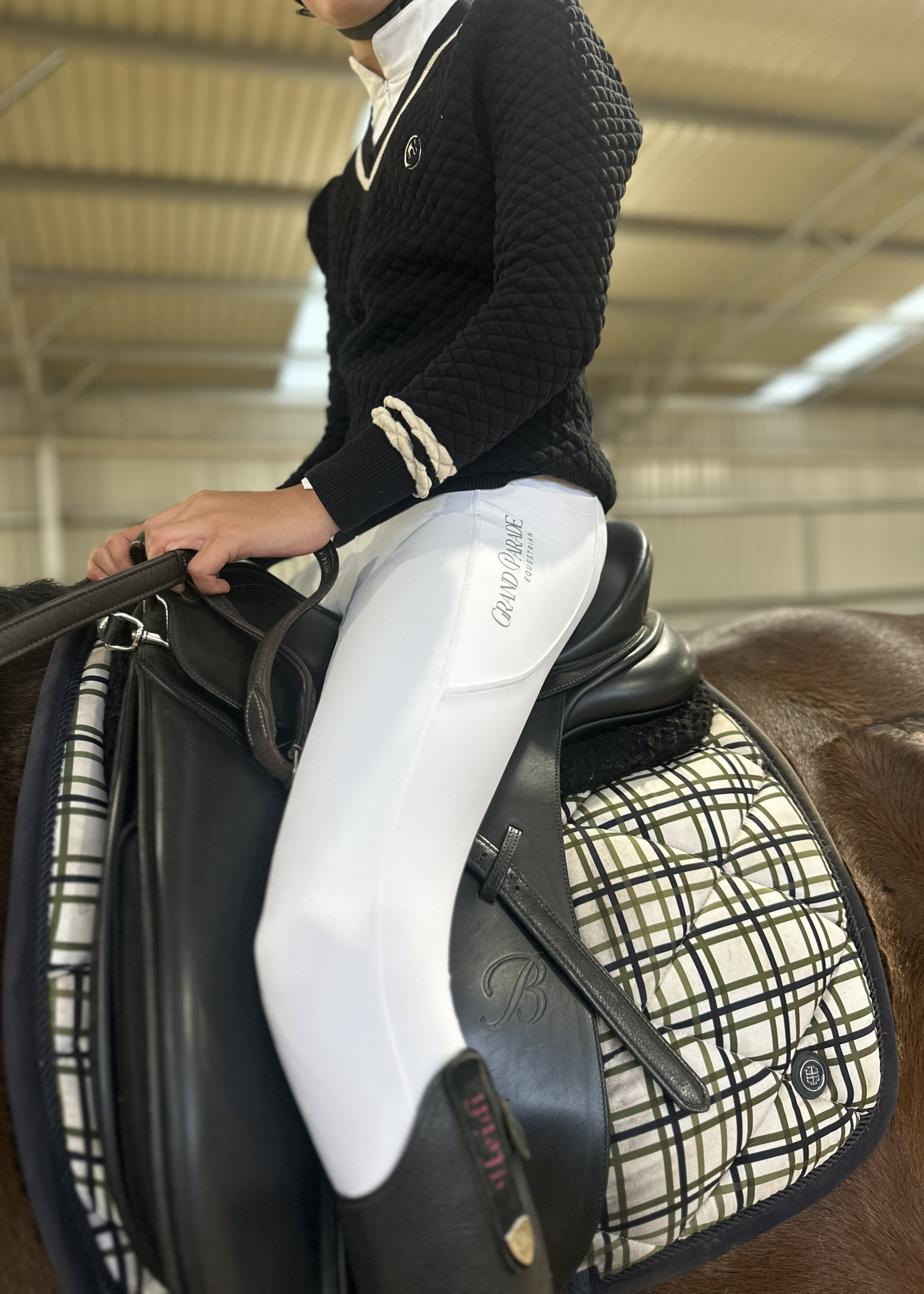 A person in equestrian attire, wearing a black knit sweater and white riding breeches with 'Grand Parade Equestrian' printed on the thigh, seated on a horse with a black leather saddle and a cream-and-black plaid saddle pad inside an indoor riding arena.
