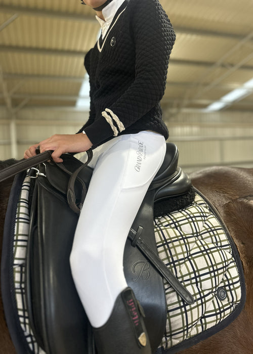 A person in equestrian attire, wearing a black knit sweater and white riding breeches with 'Grand Parade Equestrian' printed on the thigh, seated on a horse with a black leather saddle and a cream-and-black plaid saddle pad inside an indoor riding arena.