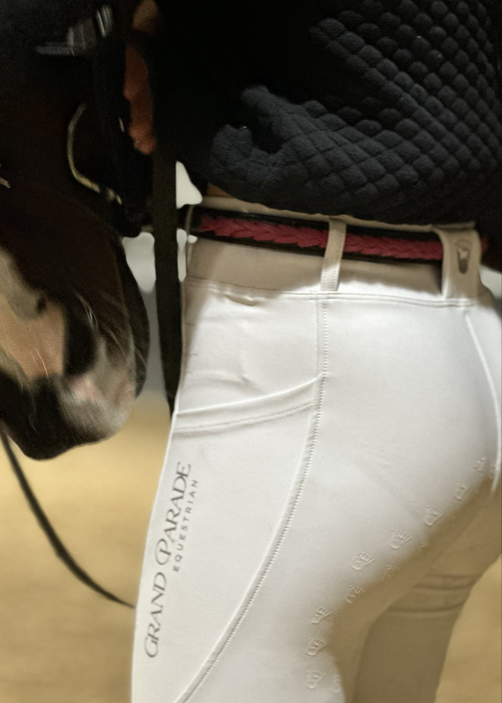 Close-up of a person wearing white equestrian riding breeches with 'GRAND PARADE EQUESTRIAN' printed vertically on the side, along with a dark quilted top and a horse's head partially visible on the left.