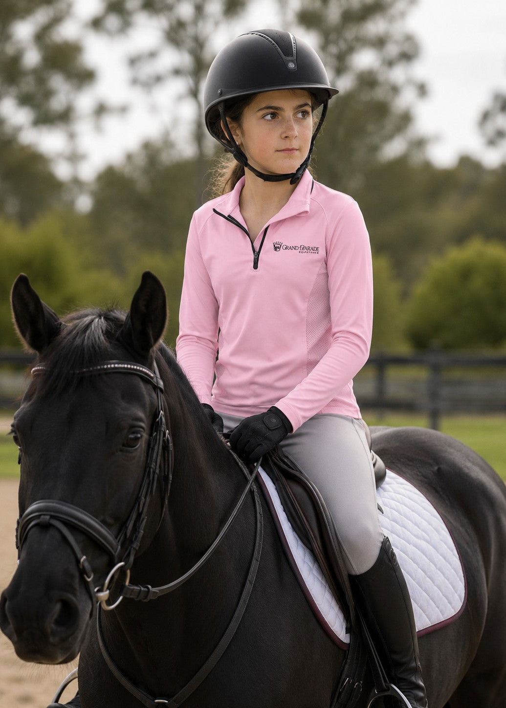 A young girl riding a black horse outdoors, wearing a pink long-sleeve equestrian shirt, light gray breeches, black riding boots, and a black riding helmet. The shirt has a visible 'Grand Parade Equestrian' logo on the left chest.