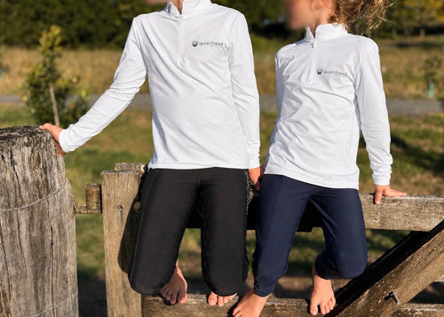 Two people outdoors wearing matching white long-sleeve quarter-zip Grand Parade Equestrian training tops with a small dark logo on the left chest, paired with black and navy pants.