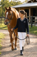 A young girl in equestrian attire, wearing a navy blue sweater with a white V-neck and white trim, white breeches, and black riding boots, walks alongside a brown horse on a dirt path in front of a stable.